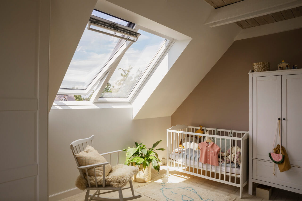 Quarto de bebê no sótão com janelas de telhado VELUX, cadeira de balanço e berço branco.