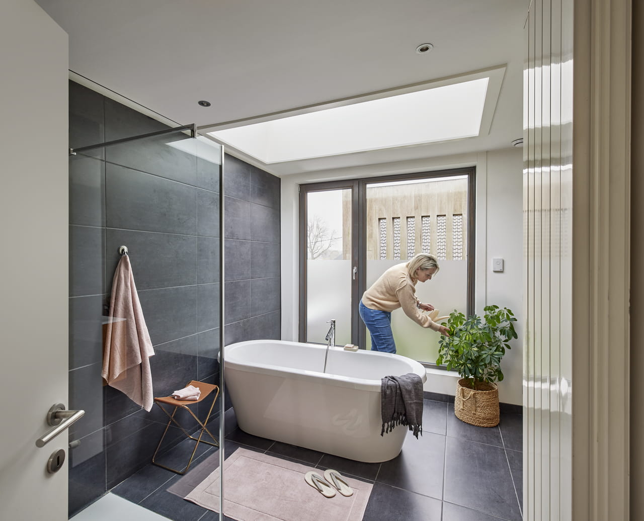 Moderne badeværelse med VELUX ovenlysvindue, fritstående badekar og person, der passer en plante.