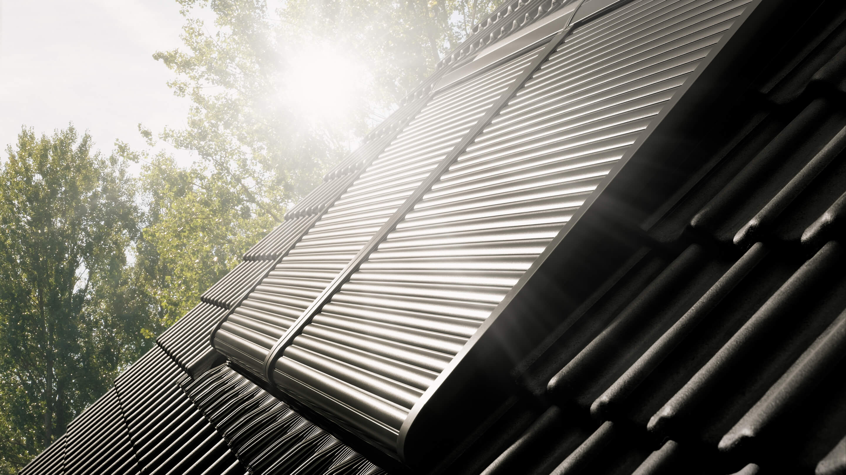 Nahaufnahme eines VELUX Dachflächenfensters mit metallenen Rollläden auf einem geneigten Dach, in Sonnenlicht getaucht.