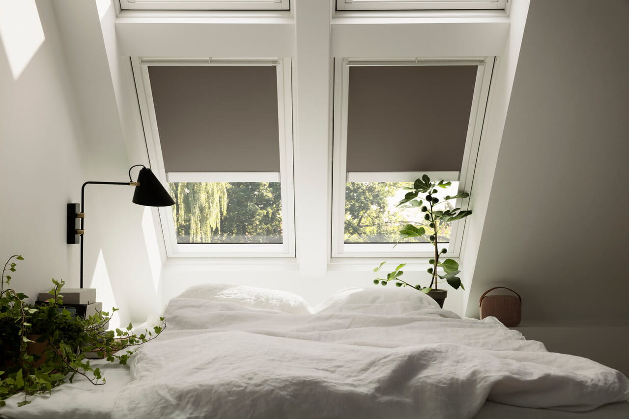 Gemütliches Schlafzimmer mit VELUX Dachflächenfenstern, ungemachtem Bett, Nachttischlampe und Pflanzen.