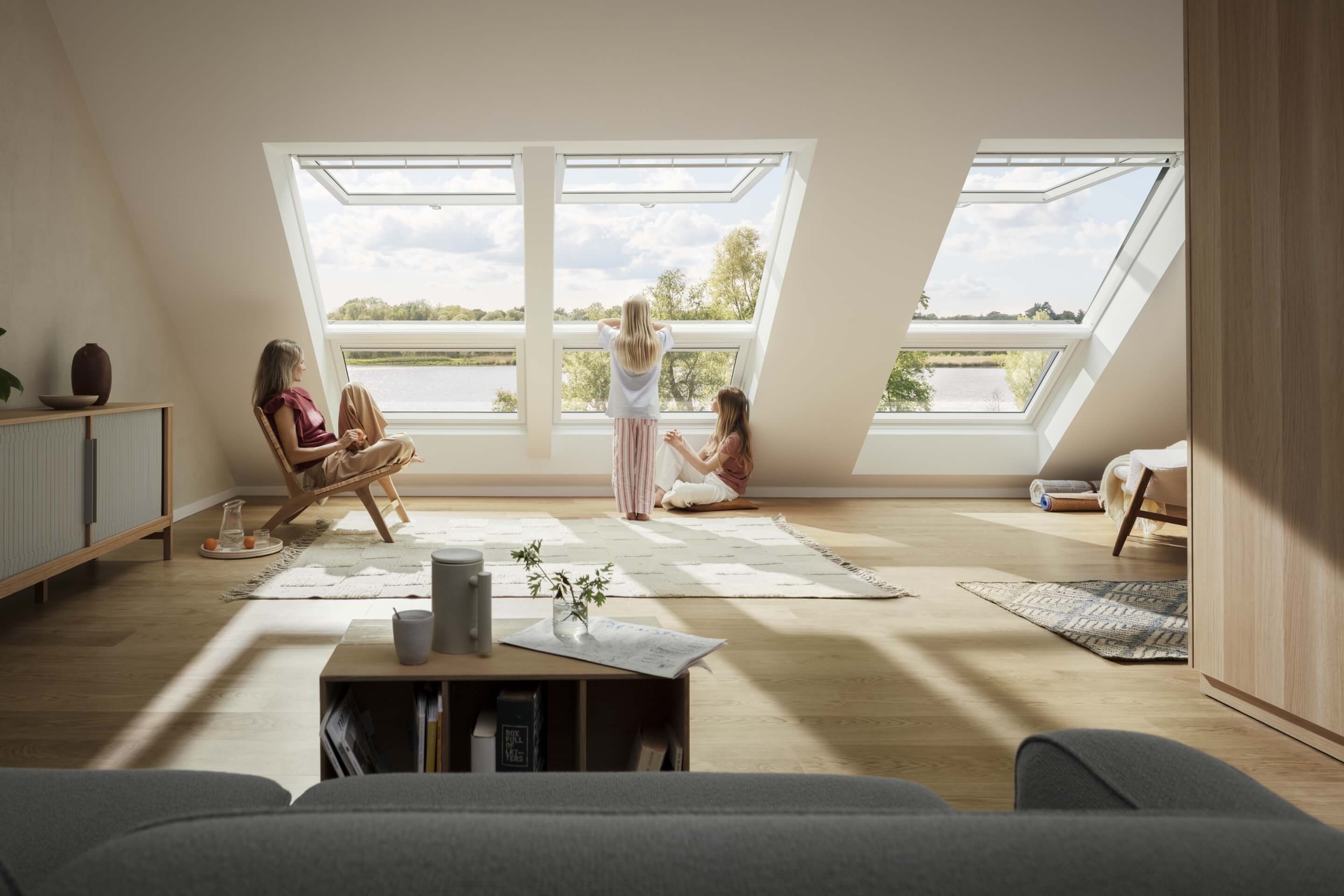 Wohnzimmer im Dachboden mit VELUX Dachflächenfenstern mit Blick auf eine Wasserlandschaft, wo eine Frau und Kinder den Raum genießen.