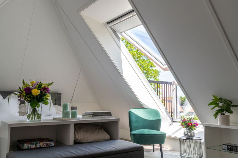 Schlafzimmer im Dachboden mit Bett, grünem Stuhl und geöffnetem VELUX Dachflächenfenster mit Blick auf den blauen Himmel.