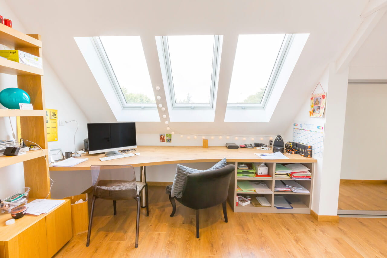 Dachboden-Heimbüro mit hölzernem Schreibtisch, VELUX Dachflächenfenster und gemütlicher Sitzgelegenheit.