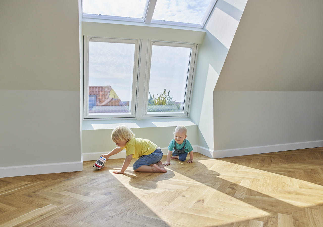 Kinder spielen in einem sonnendurchfluteten Raum mit VELUX Dachflächenfenstern und hölzernem Boden.