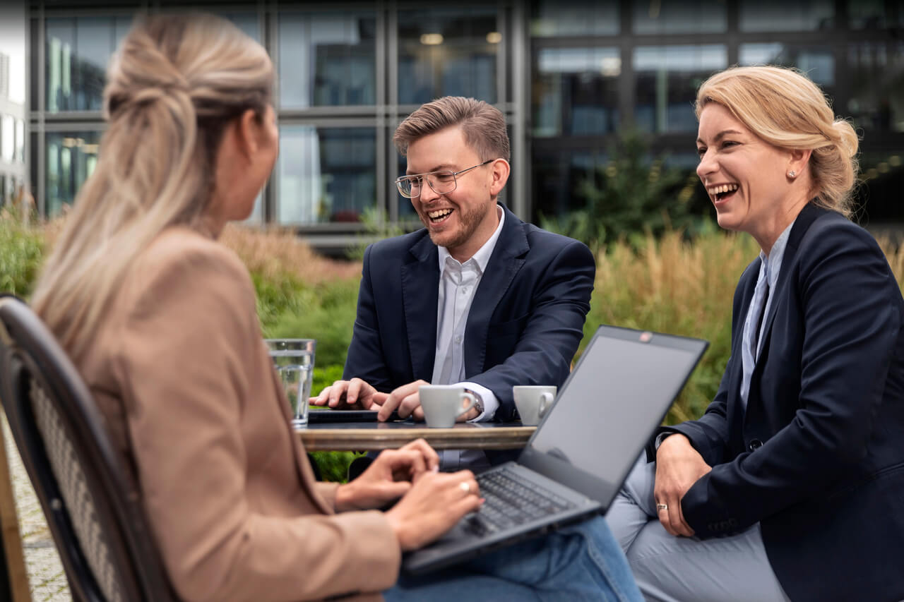 Profis bei einem Outdoor-Meeting mit Laptops an einem Tisch, Bürogebäude im Hintergrund.