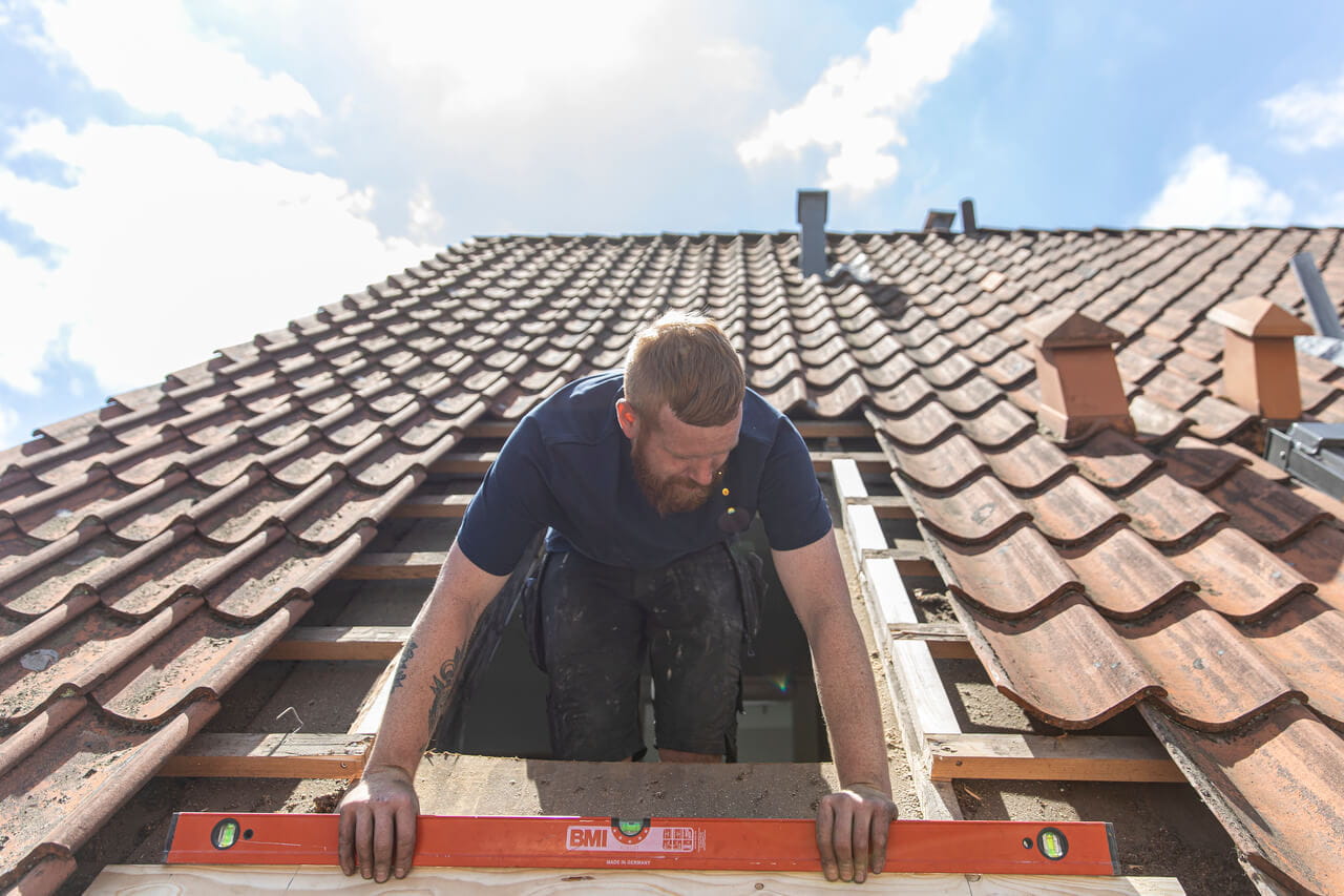 Werknemer die een VELUX dakvenster installeert op een betegeld dak met een heldere lucht.