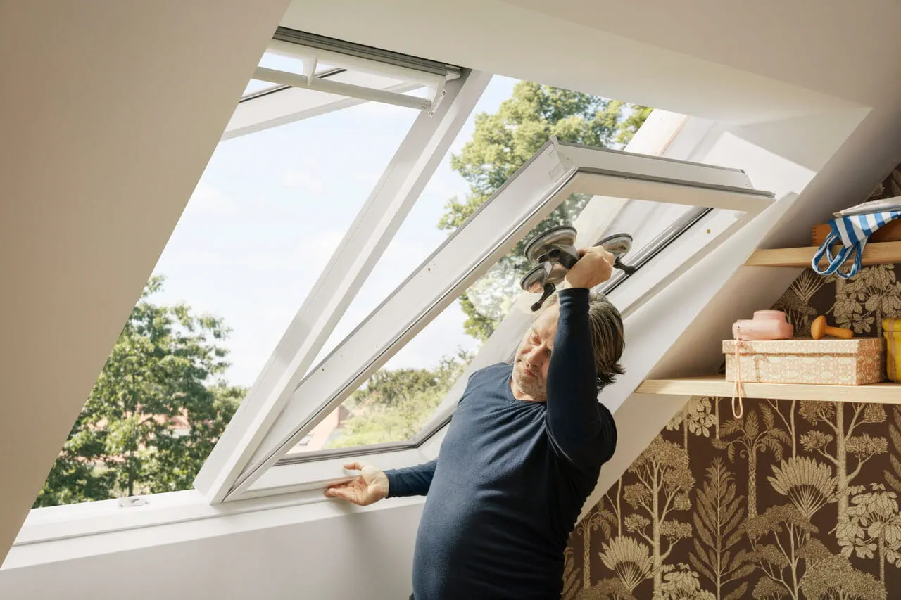 Pessoa instalando uma janela de telhado VELUX em um quarto de sótão iluminado, com vista para árvores.