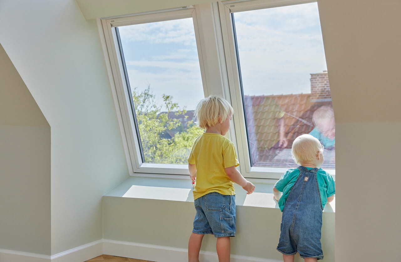 Des enfants regardent par une fenêtre de toit VELUX ouverte dans une chambre de grenier baignée de soleil.