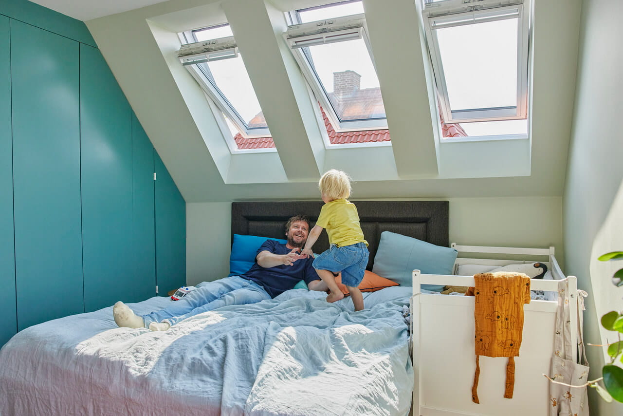 Kind speelt op een bed in een zonovergoten zolder slaapkamer met VELUX dakvensters.