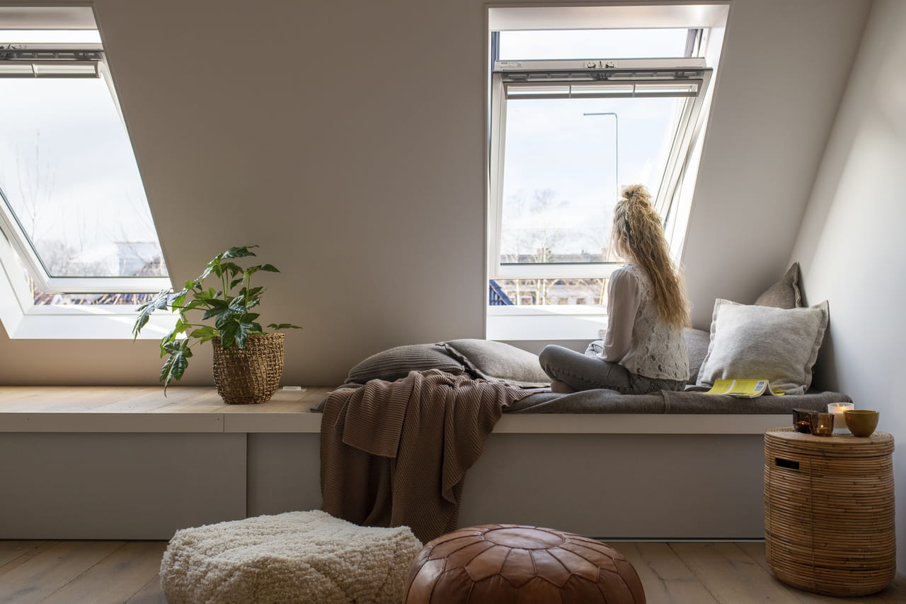 Vrouw ontspant in leeshoek op zolder bij VELUX dakvenster, met plant en gezellige decoratie.