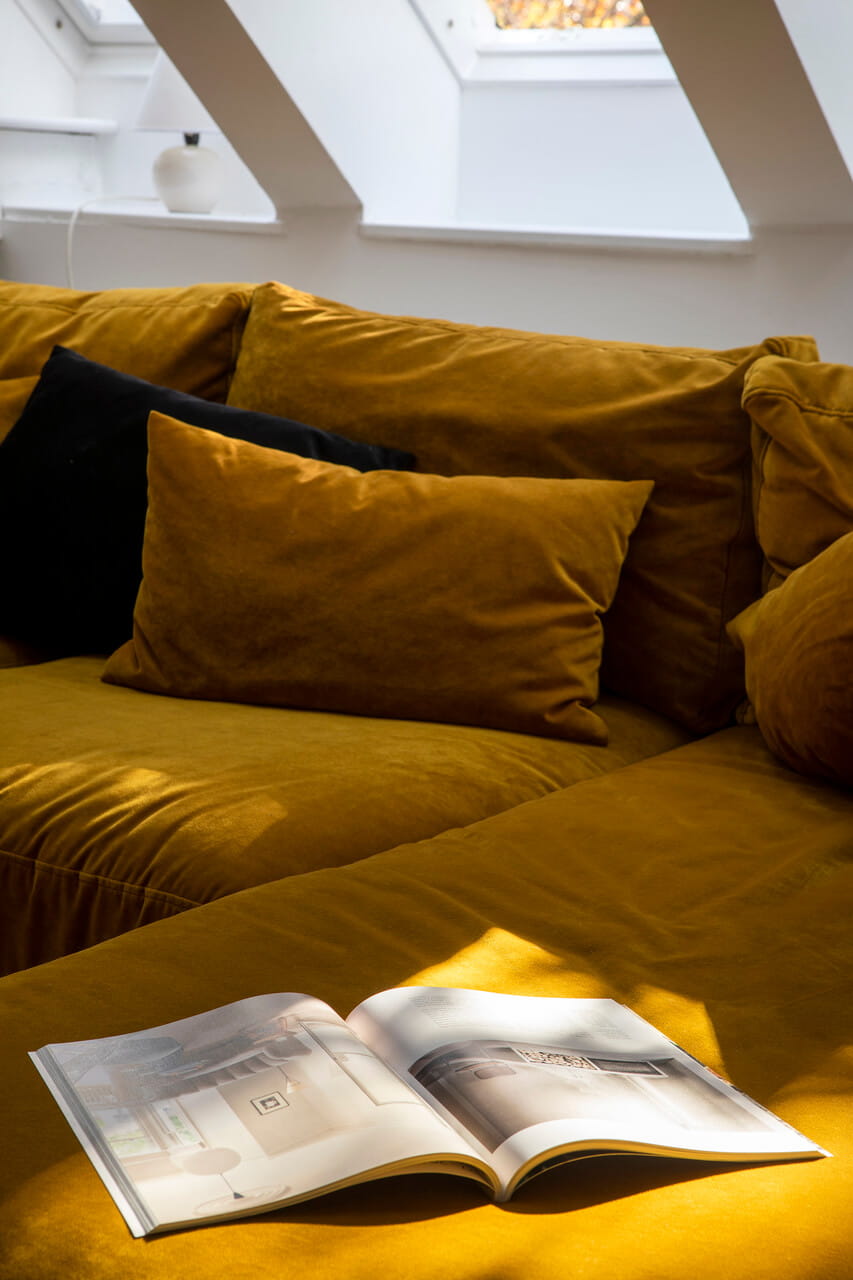 Canapé moutarde avec coussins sous une fenêtre de toit VELUX dans un coin douillet de grenier avec un magazine ouvert.