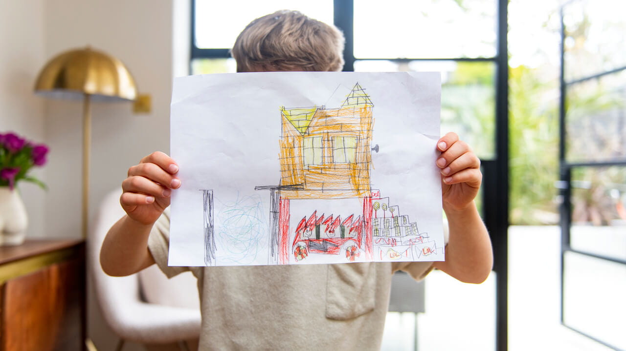 Child holding up a drawing of a house with a red car.