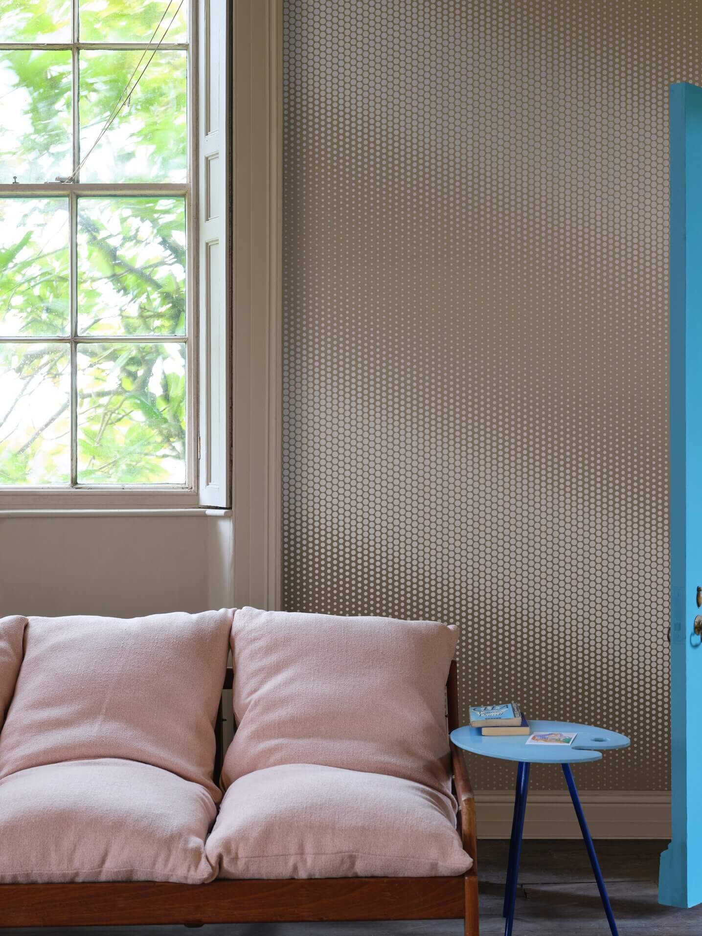 Sonnige Ecke mit rosa Kissen, gemusterter Wand und blauem Tisch neben einem großen Fenster.