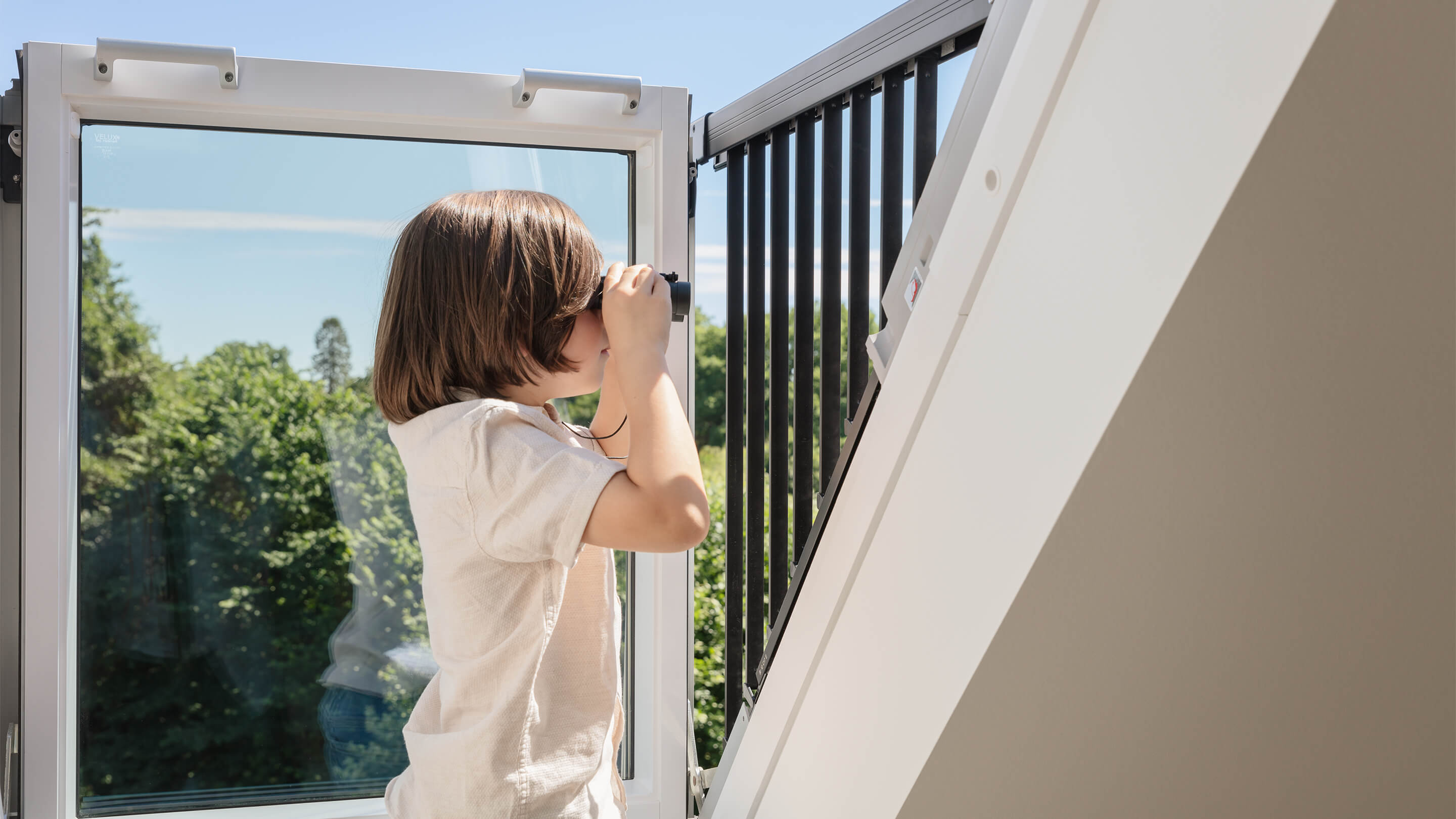 Kind benutzt ein Fernglas bei einem VELUX Dachflächenfenster mit Blick auf grüne Bäume.