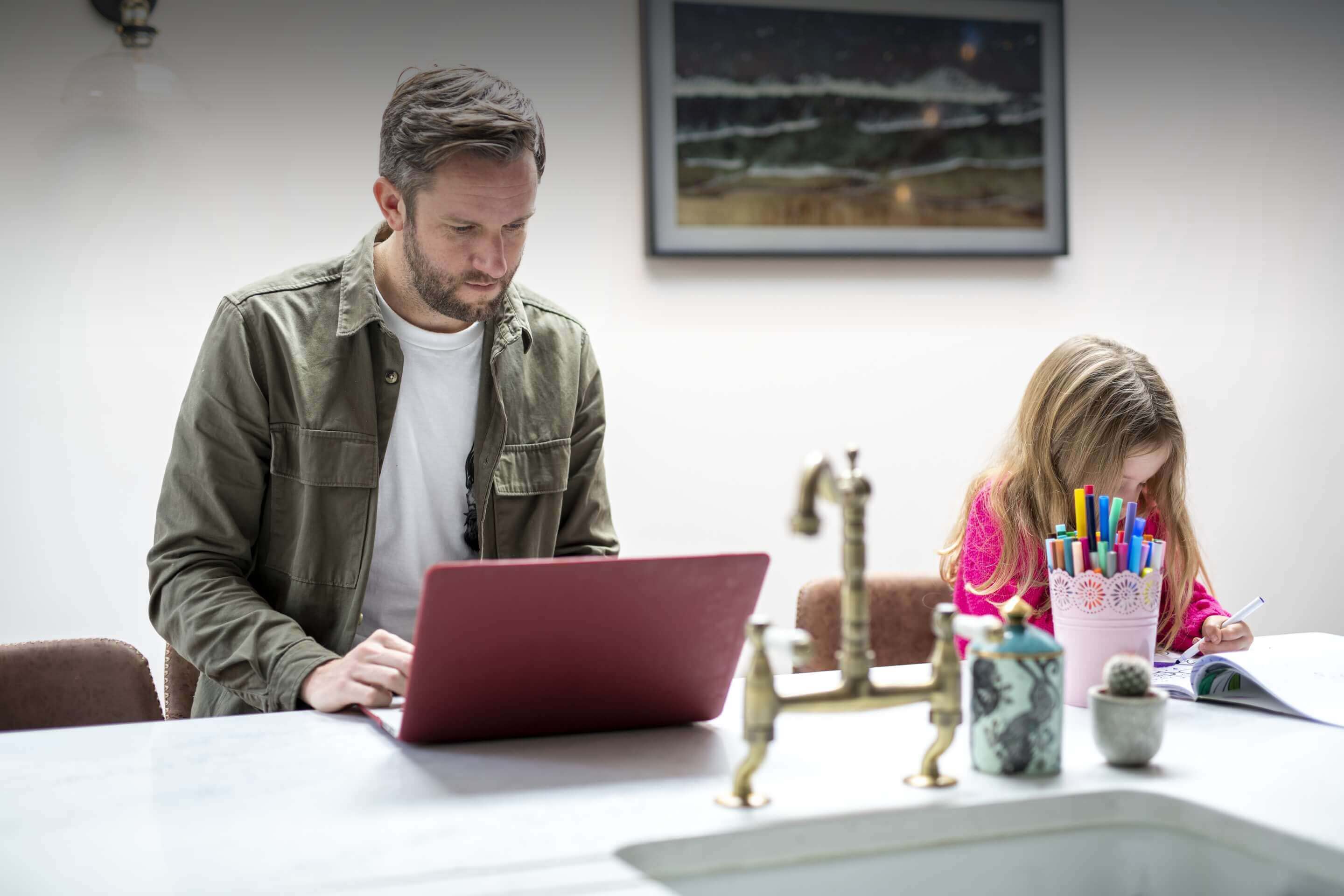 Person am Laptop und Kind beim Ausmalen in einem hellen, modernen Heimbüro.