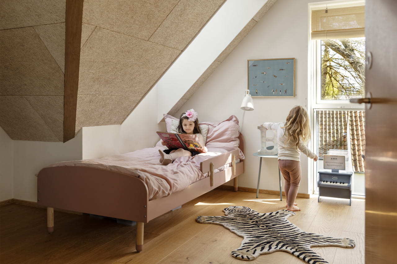 Schlafzimmer im Dachboden mit rosa Bettwäsche, hölzernen Möbeln, Tigerteppich und VELUX Dachflächenfenster.