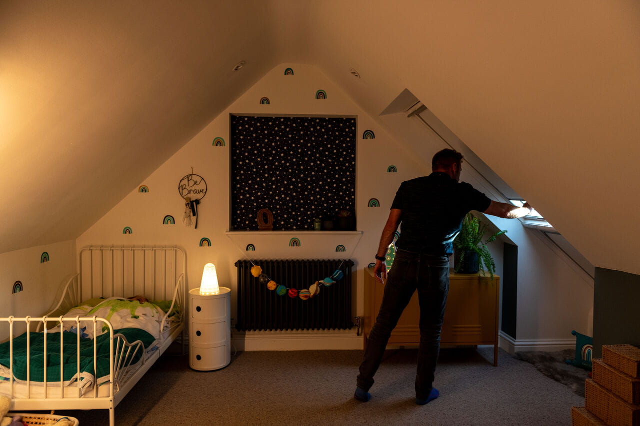 Mann justiert VELUX Dachflächenfenster in einem gemütlichen Kinderzimmer auf dem Dachboden mit warmer Beleuchtung und Dekor.